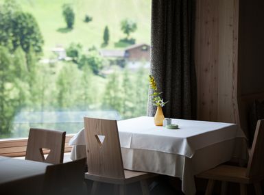 Eine elegante weiße Tischdecke liegt über einem gedeckten Esstisch vor einem großen Fenster mit Panoramablick auf die umliegenden Berge.