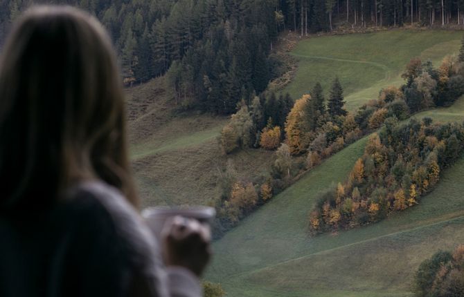 Frau blickt mit einer Tasse in der Hand auf die herbstlich gefärbten Wiesen und Wälder im Ahrntal